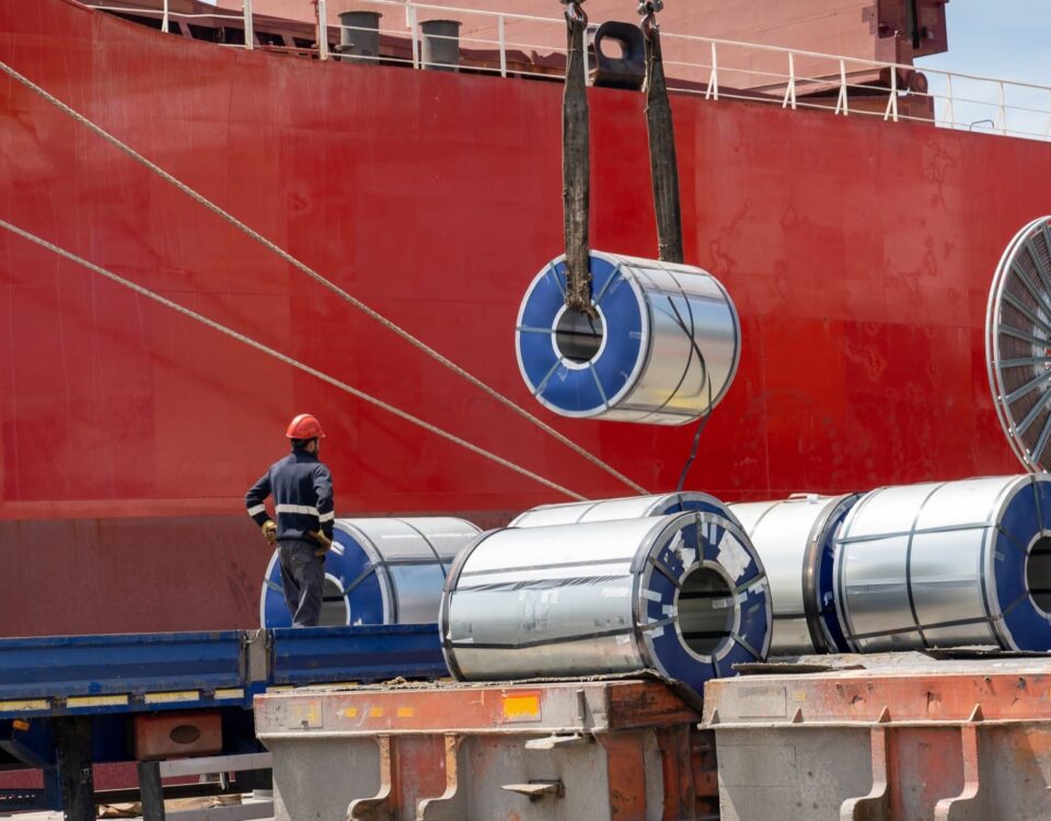 Packaged coils, galvanized steel and large tin rolls are loaded onto the ship by crane from heavy duty trucks for shipment.